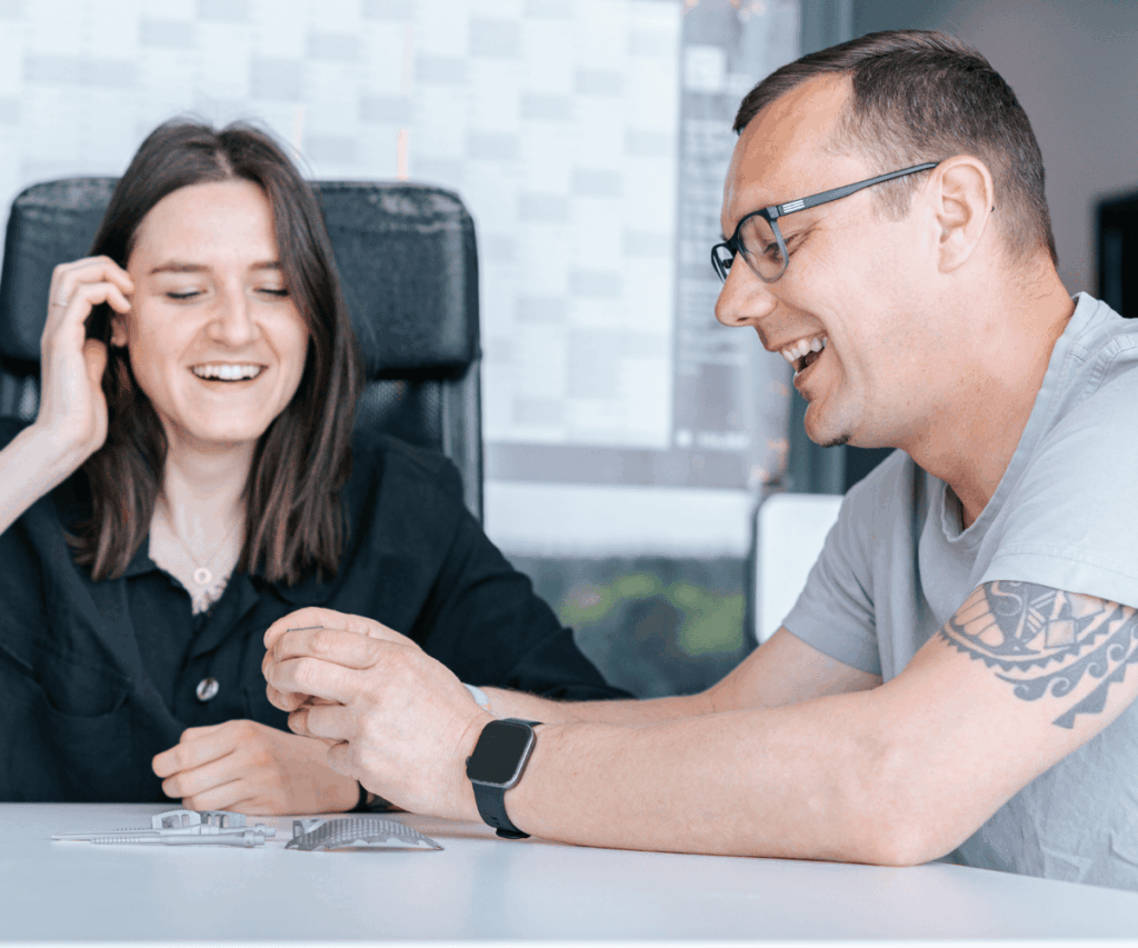 Ein Mitarbeitender von Laser Add Concept berät einen Kunden am Tisch und zeigt Metallbauteile aus dem 3D Metalldruck als Anschauungsbeispiele