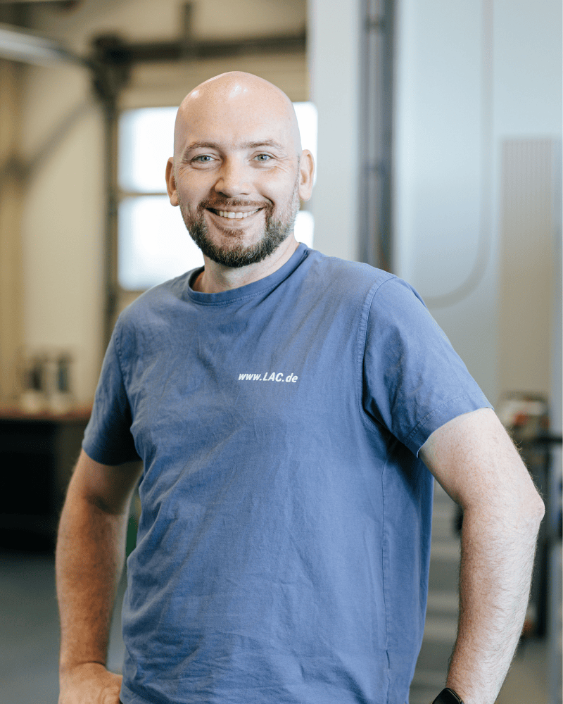 Ein Produktionsmitarbeiter von Laser Add Concept steht in Arbeitskleidung in der Fertigungshalle und schaut lächelnd mit Händen an der Hüfte in die Kamera
