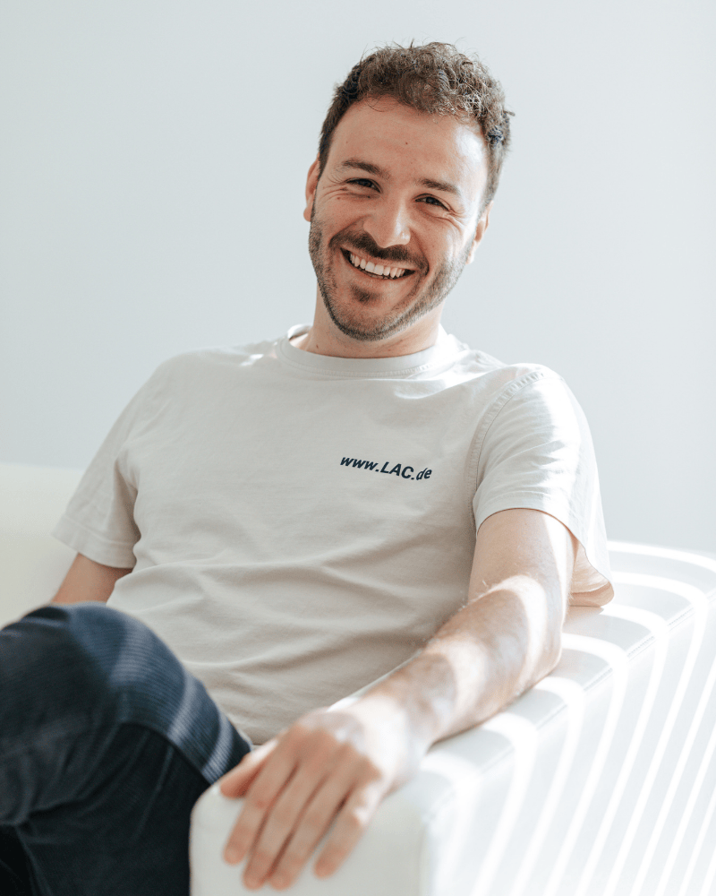 Konstantinos Lagaris, technischer Berater bei Laser Add Concept, sitzt lächelnd auf der Couch und trägt ein T-Shirt des Unternehmens